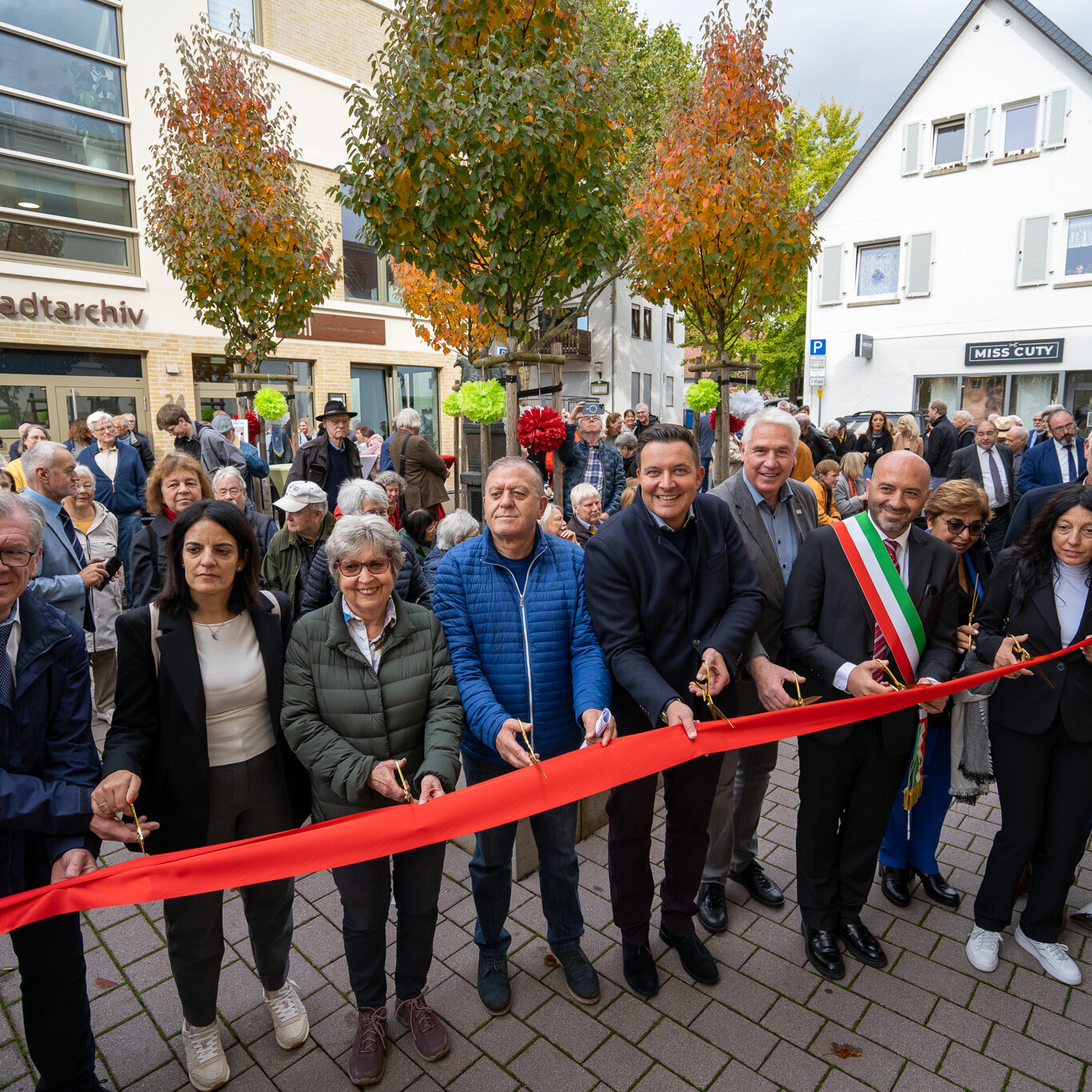 Stadtverordnetenvorsteher Andreas Hegeler (links), 1. Vorsitzende des Förderkreis Hofheimer Städtepartnerschaften Ingrid Bender (3. von links), Bürgermeister Christian Vogt (Mitte), Dezernent für Städtepartnerschaften Matthias Hees (5. von rechts), Buccinos Bürgermeister Pasquale Freda (4. von rechts) und Erster Stadtrat Daniel Phillip (rechts) mit Mitgliedern der Delegation aus Buccino beim Durschneiden des roten Bandes zur Eröffnung des Buccino Platzes.